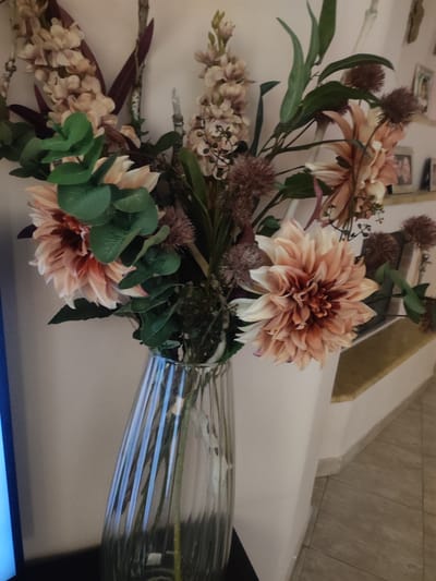 The image shows a close-up of a bouquet of artificial flowers in a clear, ribbed glass vase. The bouquet contains large, peach-colored dahlia-like flowers, smaller pinkish floral clusters, and dark green foliage. There are also some spiky, dark brown filler flowers. The vase is filled with water and the stems of the flowers are visible. The background is a plain, light-colored wall, with a hint of shelving and framed pictures visible to the right. The floor appears to be tiled. The image is focused on the flowers and vase, with the background slightly out of focus. This scene is not located in the city of Trikala, Greece.