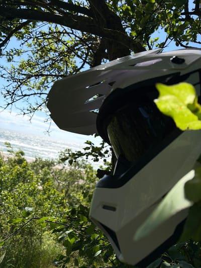 The image shows a white motocross helmet resting amongst green foliage. The helmet is positioned in the foreground, partially obscuring the background which features a bright blue sky with some scattered clouds. Beyond the foliage and sky, a body of water, possibly the sea or a large lake, is visible, with white waves suggesting it is somewhat choppy. The location is Povarovka, Russia.