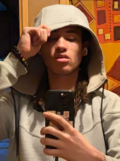 A young male individual is depicted in what appears to be an indoor setting, possibly in Orsay, France, although the specific location within the city is not visually discernible. He is positioned centrally, facing the camera directly, holding a smartphone in his right hand, suggesting a self-portrait. His left hand is raised, grasping the front edge of a light grey hooded sweatshirt, with the hood pulled up over his head. His eyes are downcast.

The subject has dark, curly hair, possibly dreadlocks, visible beneath the hood. He wears a white wireless earbud in his right ear. On his left wrist is a bracelet composed of dark beads interspersed with several brightly colored, multifaceted beads (red, green, yellow). A thin silver chain bracelet is visible on his right wrist.

The smartphone held in his right hand is dark-colored, featuring a multi-lens camera array and decorative stickers or elements on its back panel.

The background is out of focus, displaying warm tones. To the right, an abstract pattern on a wall features geometric shapes in orange, red, and dark brown. To the left, a darker, indistinct area transitions into a vertical wooden surface. The lighting is warm and appears to be artificial.