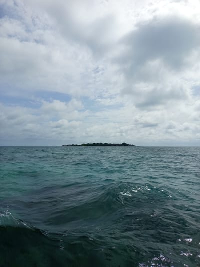A wide shot captures a serene ocean scene with a distant island under a cloudy sky. The foreground is dominated by dark blue-green water, with small waves creating ripples and reflecting sunlight. In the middle ground, the ocean continues, with a hazy horizon separating it from the sky. The background features a long, narrow island covered in lush green trees and dotted with a few buildings. The sky above is a mix of white and gray clouds, with patches of blue sky visible. The overall impression is one of a calm, natural environment.