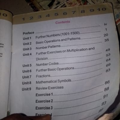 The image shows a table of contents from a book. The book appears to be a mathematics textbook, as indicated by the titles of the units such as "Further Numbers," "Basic Operations," "Fractions," and "Mathematical Symbols." The table of contents lists units 1 through 9, along with page numbers. The book is open to a page with the heading "Contents" printed in a pinkish-purple font. The page is bordered by a thick, light brown strip at the top, which has numbers 1 through 10 printed on it. The lighting in the image suggests it was taken indoors, possibly in a dimly lit room, with the flash from the camera illuminating the page. The book is lying flat, and a portion of a hand is visible at the bottom of the image, suggesting someone is holding or browsing the book. The background is a textured surface, likely a table or floor. There is no information in the image to suggest the location is in Jauro Bamvo, Nigeria.