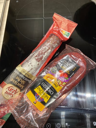 Two packaged dried sausages are depicted resting on a black, glossy induction stovetop in Bonnieux, France. The stovetop features four marked cooking zones and a partial control panel at the bottom right, displaying symbols for operation (e.g., minus, plus, power).
The upper-left object is an elongated, cylindrical *saucisson sec* (dried sausage), wrapped in clear plastic and a red paper label. The label identifies it as "Saint Azay Saucisse Sèche Qualité Supérieure Pur Porc" and displays a Nutri-Score rating, indicating a B or C. The sausage itself exhibits a characteristic white mold coating.
Below and partially to the right of the *saucisson* is a curved, reddish chorizo sausage, encased in clear plastic netting with a black and yellow label. This label prominently states "CESAR MORONI CHORIZO Doux Recette Originale" and indicates a net weight of 200g, along with claims of being "N°1" and having "-30% Mat. Grasse" (30% less fat). No individuals are present, and the objects are stationary on the cooking surface.