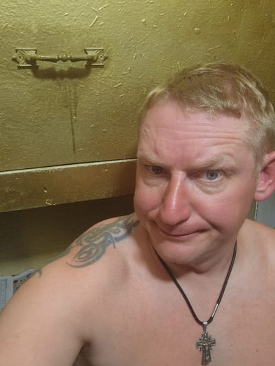 A shirtless man with blond hair and blue eyes poses for a selfie in front of a textured, golden wall. He is wearing a dark cord necklace with a silver cross pendant. A tribal tattoo is visible on his left shoulder. The background shows a brass handle and a shelf. The man appears to be in a room with a unique, possibly metallic, finish. The location is specified as Svetogorsk, Russia.