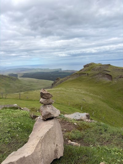 This wide-angle landscape photograph depicts a rugged, verdant terrain near Portree, United Kingdom, under a completely overcast sky. In the foreground, slightly to the left of the frame's center, a small cairn is constructed from three stacked, irregularly shaped, light grey-brown stones resting on a patch of green grass with visible soil and some low-lying clover. The largest stone forms the base, supporting two progressively smaller, more rounded stones above it.
The immediate foreground descends into a steep, grass-covered slope that gives way to a expansive valley. On the left midground, several slender, light-colored wooden fence posts are sparsely placed, delineating a boundary down the hillside. To the right, a prominent, steep, grass-covered hill or ridge features rocky outcrops and a distinct concave erosion or natural depression on its slope. The valley floor in the distant midground shows varying shades of green vegetation, including darker areas potentially indicating forests, extending towards a body of water.
In the background, a calm, light blue-grey expanse of water, presumably the sea, stretches to the horizon, bordered by a distant, undulating coastline. The entire sky is covered by a dense, uniform layer of grey stratocumulus clouds, indicating overcast weather conditions with soft, diffused light. No individuals or animals are present in the image.