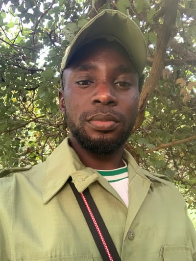 An adult male, appearing to be of African descent, is depicted in a head-and-torso portrait, facing directly towards the camera. He has dark skin, dark eyes, and a short, neatly trimmed dark beard and mustache. He wears a light olive-green collared, button-up shirt with visible shoulder epaulets and a subtle ripstop fabric pattern. Beneath this, a white t-shirt with green and white horizontal stripes around the collar is visible. A light olive-green baseball cap is worn on his head. A black strap featuring a red and white zig-zag pattern is slung diagonally across his chest. The background consists of dense green foliage, including visible tree branches and leaves, indicating an outdoor, vegetated environment under natural daylight. This scene is located in Kubi A, Nigeria.