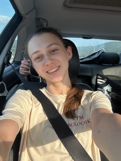 A young woman with light brown hair and blue-green eyes is taking a selfie in the driver's seat of a car. She is wearing a cream-colored t-shirt with red lettering that reads "I'M BULOCHK". She is smiling and her seatbelt is fastened. The interior of the car is visible, including the dashboard and part of the passenger seat. Outside the car window, a body of water and trees can be seen, suggesting a scenic location. The lighting indicates it is daytime. The orientation of the image suggests the photo was taken while the woman was driving or in the passenger seat, looking towards the driver's side. The photo appears to have been taken in Bürgstadt, Germany.