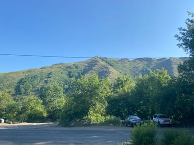 The image depicts a scenic view of a mountain range covered in lush green trees under a clear blue sky. In the foreground, there is a paved road with white dashed lines. To the right of the road, two SUVs are parked. The one on the left is a dark-colored SUV, and the one on the right is a silver SUV. Behind the vehicles, a dense line of trees partially obscures the base of the mountain. The mountain itself is steep and covered with a mixture of lighter green shrubs and darker green trees, with visible textures and contours. A thin black wire stretches horizontally across the upper portion of the frame, separating the sky from the landscape. The overall scene suggests a natural, possibly rural or mountainous area on the outskirts of a city. Given the context of Oskemen, Kazakhstan, this could be a view from a road leading into or out of the city, showcasing its proximity to natural landscapes.