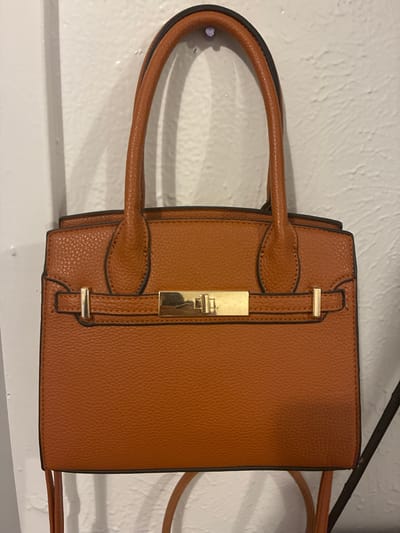 A brown, pebbled leather handbag with a gold clasp and gold buckles hangs on a white textured wall in Balch Springs, USA. The handbag has two top handles and appears to be empty. The lighting is dim, casting shadows on the wall behind the bag. The image is a close-up shot, focusing solely on the handbag and its immediate surroundings.