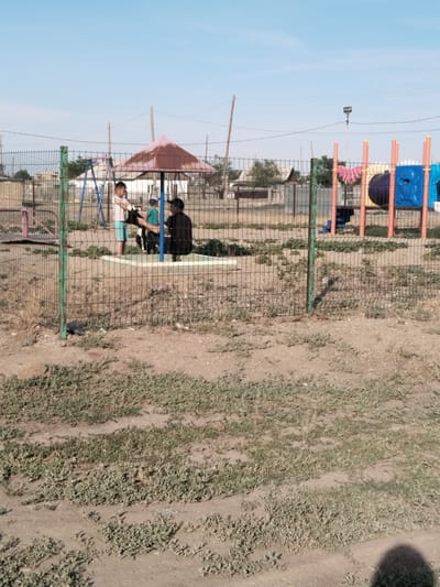 In the city of Ақадыр, Kazakhstan, a playground scene unfolds under a clear blue sky. A chain-link fence, painted green at the posts, separates the foreground from the playground. Within the fenced area, a red and pink hexagonal umbrella provides shade. Underneath the umbrella, a boy in a striped shirt and blue shorts is handing something to a man who is crouching. A smaller child in a turquoise shirt and a blue cap is also present near the umbrella.

To the left of the umbrella, a blue swing set stands, and in the background, several structures and a tunnel playground with orange and blue elements are visible. The ground within the playground appears to be a mix of dirt and sparse vegetation. The area outside the fence, in the foreground, is covered with dry, patchy grass. Utility poles and power lines stretch across the sky, indicating the presence of infrastructure in the vicinity. The overall setting suggests a public space, likely a community park or schoolyard, in a semi-arid environment.