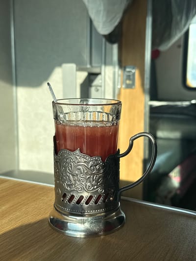A clear, faceted glass containing a reddish-orange liquid, consistent with a beverage, is centrally positioned. A small silver spoon rests vertically within the liquid. The glass is held by an ornate silver-colored *podstakannik* featuring embossed floral and scrollwork designs, and a sturdy handle on the right. This assembly rests on a light-colored wooden surface with a visible grain, likely a table or tray.

Direct sunlight illuminates the scene from the left, casting sharp shadows from the glass and spoon onto the wooden surface and the background. The background is out of focus, revealing the interior of what appears to be a train carriage. Visible elements include a bright, light-colored wall section on the left, a vertical wooden panel or frame to the right of center, and a blurry window or opening on the far right showing bright light. Above, two indistinct objects, possibly wrapped luggage or covers, are partially visible.

No people are present. No direct actions or interactions are depicted. The scene is static.

Regarding the specific location in Volkhov, Russia: The image contains no discernible visual markers, such as specific landmarks, unique architectural features, or identifiable signage, to forensically confirm its exact location within the city of Volkhov, Russia. The depicted environment is consistent with a generalized train interior, commonly found across Russian rail networks.