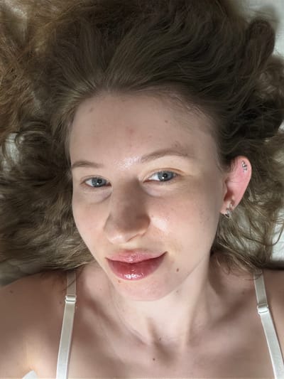 A close-up, overhead shot shows a young Caucasian woman lying down with her head tilted back and hair spread out around her. Her eyes are open and looking at the camera, and she has light makeup on, with glossy lips. She is wearing a white bra strap visible on her shoulder. The background appears to be a white sheet or bed. There are no discernible elements indicating a specific location in Dongducheon-si, South Korea, or any other city.