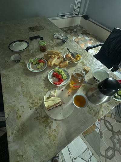An overhead, slightly angled view depicts a marble-patterned dining table set for a meal, presumably in Jauqashty, Uzbekistan. The table is laden with various food items and serving dishes, including sliced bread, a plate of sliced cucumbers and tomatoes, a plate with what appears to be a rice dish and potatoes, a jar of honey, a slice of layered cake with green frosting, and a small bowl of amber liquid, possibly tea or juice. A stainless steel carafe with a black lid, a fork, and two bowls are also present. The background shows a tiled floor and a chair with a black woven back. The lighting suggests an indoor setting, possibly during daytime or with artificial illumination.