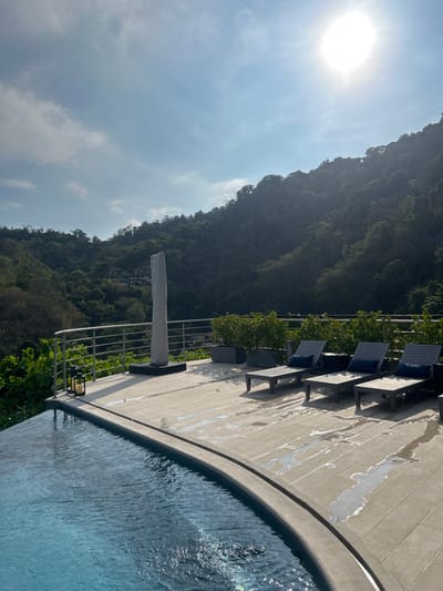 The image shows an outdoor swimming pool at a resort in Playa Herradura, Costa Rica. The coordinates (latitude: 9.658, longitude: -84.673) place the location on the Pacific coast of Costa Rica, within the resort area of Playa Herradura. The swimming pool is surrounded by a deck with three lounge chairs. The deck is made of light-colored tiles and is bordered by a metal railing. There is a large, gray, column-shaped sculpture on the deck. The background consists of lush green vegetation, including trees and shrubs, covering a hillside. The sun is shining brightly and is reflected off the water in the swimming pool. The scene suggests a luxurious resort experience.