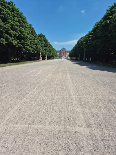 The image depicts a wide, gravel-covered pathway leading towards a grand, ornate building in the distance, likely a palace or significant historical structure. The pathway is flanked by symmetrical rows of mature, dense green trees, creating a natural avenue. Two classical statues stand on pedestals on either side of the pathway, midway down its length. The sky is a clear, bright blue with a few wispy clouds. In the middle distance, on the right side of the pathway, a single person, wearing a white shirt and dark shorts, is walking away from the camera. The architectural style of the building suggests a European Baroque or Rococo design, with a prominent central section featuring a mansard roof, arched windows, and a facade with columns and decorative elements. The scene is bathed in sunlight, casting shadows from the trees onto the pathway. The location is the grounds of Bruchsal Palace (Schloss Bruchsal) in Bruchsal, Germany.