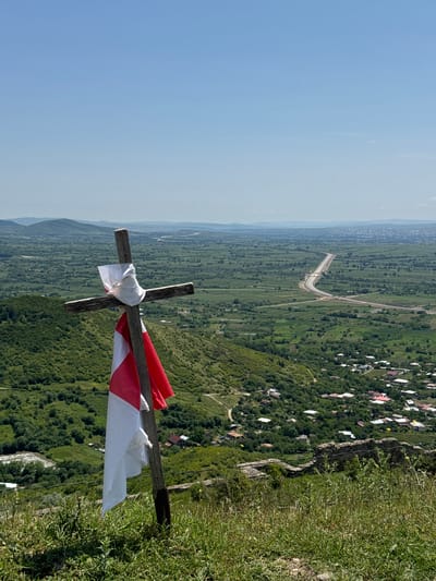 A wooden cross stands prominently in the foreground, adorned with a white cloth tied at the top and a white and red flag draped down its length. The cross is situated on a grassy hilltop overlooking a sprawling valley. The valley is a tapestry of lush green vegetation, with scattered houses forming a small village. A highway cuts through the landscape in the mid-ground, with hills receding into the distance under a clear blue sky. The scene appears to be taken from a high vantage point, suggesting a significant elevation. The image captures a serene, rural landscape in Manavi, Georgia.
