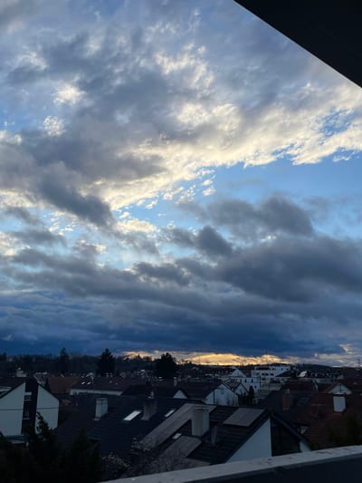 An elevated, panoramic view of a residential area in Senden, Germany, is depicted at what appears to be dawn or dusk. The sky dominates the upper two-thirds of the image, characterized by a dynamic mixture of dark, heavy stratocumulus clouds partially illuminated by direct sunlight, revealing patches of lighter blue sky. A distinct band of bright golden-orange light is visible on the horizon, primarily in the lower-middle portion of the sky, indicating the position of the sun, which is obscured by clouds.
The lower third of the image showcases a densely packed urban landscape composed predominantly of residential houses with varied rooflines. Many roofs are steeply pitched, featuring reddish-brown terracotta tiles, while others are dark grey or black, often equipped with visible skylights and solar panels. Chimneys are frequently observed protruding from the rooftops. Buildings exhibit a mix of architectural styles, generally two to three stories in height, with white or light-colored facades visible on some structures. Interspersed among the houses are various trees, some appearing evergreen (coniferous) and others deciduous, with sparse foliage. In the far distance, a treeline follows the contour of rolling hills, and a faint structure, possibly a church spire or tower, is silhouetted against the bright horizon light. The image is framed in the lower right by a dark, horizontal barrier or railing, suggesting the vantage point is from a balcony or elevated platform. No individuals or discernible activities are visible within the scene.