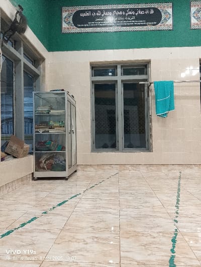 The image shows the interior of a building located at latitude -6.423, longitude 107.996 in Kecamatan Kroya, Indonesia. There is a glass-fronted cabinet with shelves that is filled with various items, including books and clothes. The cabinet is located in the corner of the room, near a window. The room has tiled floors with green painted lines running across the floor. There are two windows on the far wall and a teal colored towel is hanging on a clothes rack. The walls are tiled and cream colored. There is a ceiling fan in the top left corner of the image. The room is empty of people. The scene is brightly lit and appears clean and well-maintained.