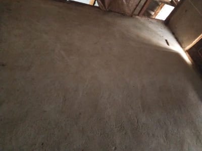 The image shows the interior of a room under construction. The walls are rough, unfinished concrete. In the upper right corner, wooden beams and rafters of the ceiling are visible, with gaps allowing in natural light. The lighting suggests it is daytime, with sunlight casting shadows on the wall, creating a contrast between illuminated and darker areas. A small, dark, elongated object protrudes from the wall in the upper left quadrant, possibly a nail or a piece of wire. The overall scene depicts an early stage of building or renovation, with a focus on the raw materials and structural elements of the room. The setting appears to be Gindin Dutse, Nigeria, implying a rural or developing area context. There is no visible text in the image.