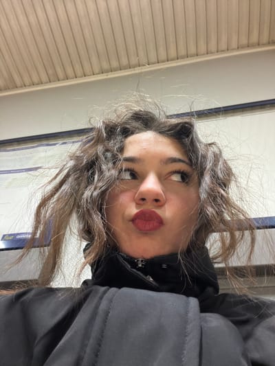 A young woman poses with pursed lips, her gaze directed upwards. She has wavy, light brown hair and wears a black jacket. The background includes a wall with horizontal lines and a digital screen displaying text and graphic elements. The overall composition suggests a candid or selfie-style photograph, with a casual and slightly tilted perspective.
