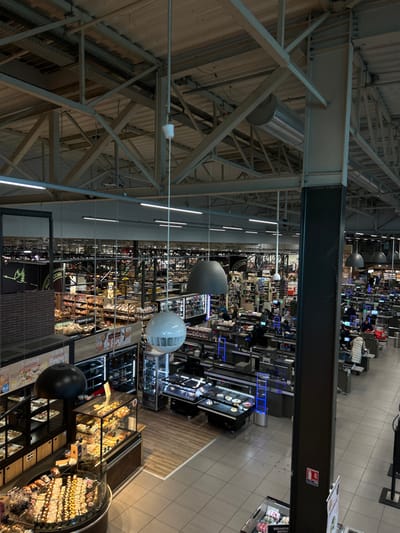 An overhead view captures a large, active supermarket interior, identified by signage as located in Eschau, France, likely within a "Vallée de l'Eschau" establishment. The expansive space features a high ceiling with exposed metal structural trusses, cylindrical air ducts, and rows of fluorescent strip lighting. Several large pendant lights, including dome-shaped grey fixtures and a white spherical one with vents, hang throughout the store.
In the foreground and midground, a substantial bakery and patisserie section is visible on the left, featuring multiple glass display cases filled with various pastries, breads, and baked goods. A prominent round display case is at the bottom left, showcasing numerous small items. Signs in French, such as "la fournil," are present. Adjacent to this, and extending towards the right, is a busy area containing numerous checkout lanes, some appearing to be self-service, marked by blue illuminated barriers.
Approximately 15-20 individuals are present. Several people are engaged in transactions at the checkout lanes, with some standing at the counters and others pushing shopping carts. Staff members are visible operating registers. Other individuals are dispersed throughout the background aisles, browsing products on shelves. Refrigerated display units containing packaged items are positioned near the checkout area. The flooring consists primarily of light grey tiles, with a section of wood-look flooring near the bakery. The background reveals extensive, well-stocked grocery aisles with various products arranged on shelves, extending deep into the store.