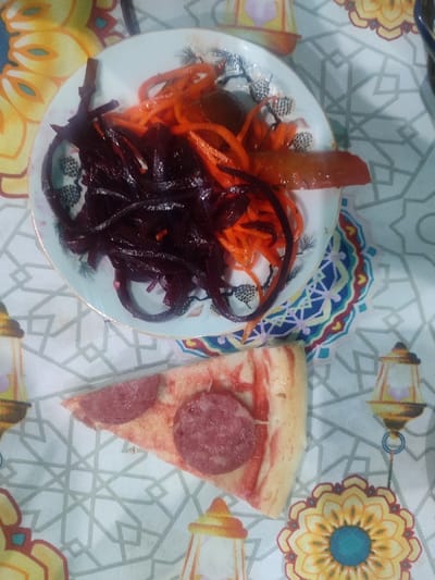 An overhead view captures a meal laid out on a decorative white tablecloth in Rishtan, Uzbekistan. A circular white plate, edged with a subtle gold rim and a dark, abstract sprig pattern, is positioned centrally in the upper half of the frame. The plate holds a serving of shredded, dark reddish-purple vegetables (consistent with beetroot) on the left side, and a portion of bright orange shredded vegetables (consistent with carrots) on the right. Several elongated, light orange-brown vegetable pieces are also present on the right side of the plate, partially obscured by the shredded carrots. Below the plate, a triangular slice of pizza rests directly on the tablecloth. This pizza slice features a pale crust, faint red sauce streaks on its surface, and two distinct round slices of dark pink salami or pepperoni as toppings. The tablecloth itself displays a light grey geometric grid pattern over a white background, interspersed with colorful, stylized motifs of yellow lanterns and blue, yellow, and red mandalas or floral designs. No individuals are visible.
