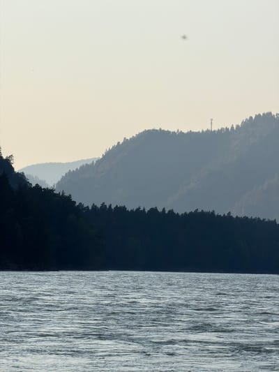 The image depicts a serene landscape of mountains, a forest, and a body of water. In the foreground, a wide river with rippling water flows, reflecting the subdued light. The midground features a dense, dark forest covering the lower slopes of rolling hills. Further in the background, layers of hazy, blue-hued mountains recede into the distance, their peaks softened by the atmospheric perspective. Atop one of the distant mountain ridges, a solitary cell tower is visible, a man-made structure contrasting with the natural environment. The sky is a pale, overcast grey, with a faint reddish-orange object suspended in the upper portion of the frame, possibly a distant balloon or drone. The scene evokes a sense of tranquility and vastness, characteristic of natural landscapes. The location is identified as Anos, Russia.
