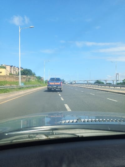 The image captures a daytime scene from inside a moving vehicle, looking forward through the windshield. The dashboard occupies the bottom portion of the frame, with reflections visible on the glass above it. Ahead, a blue pickup truck, possibly a Ford Ranger, is driving in the same direction on a multi-lane asphalt road. The truck's tailgate features yellow and red reflective diagonal stripes, and its license plate "MN-24-95" is clearly visible.

The road features white dashed lines separating the lanes. To the left, a grassy embankment rises, topped by a metal guardrail, scattered green trees, and several multi-story buildings. Tall streetlights line both sides of the road, extending into the distance. The right side of the road appears wider, with more lanes and a concrete barrier separating it from a lower section or another part of the road infrastructure.

In the far background, a prominent white cable-stayed bridge spans across what appears to be a body of water or a wide river, with its distinctive towers and cables visible under a bright, clear blue sky with a few faint white clouds. The weather is sunny and clear, suggesting a pleasant day. The overall scene depicts travel on a major highway or approach road leading to a large bridge, consistent with the Maputo-Katembe Bridge in Maputo, Mozambique.