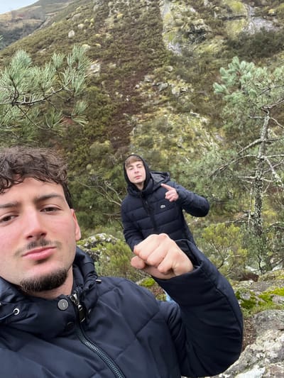 A daytime photograph captures two young adult males in a rugged, vegetated hillside environment within Ribeira de Pena, Portugal. The foreground subject, a male with curly brown hair and a short beard, wears a dark navy puffer jacket and looks directly into the camera. His right arm is bent, with his fist clenched and elevated towards the viewer. Positioned behind and to the right, a second male with lighter hair wears a dark navy hooded puffer jacket with a white crocodile logo visible on the left chest. He also looks towards the camera, extending his right arm with the index finger pointed upwards. The background reveals a steep, rocky slope densely covered with green and reddish-brown scrub vegetation and scattered pine trees. Pine needles are visible in the upper left and right portions of the frame. The sky appears overcast, providing diffused natural lighting.