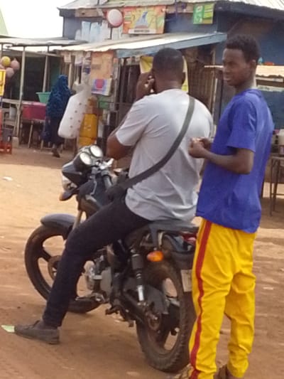 The image shows two men and a parked motorcycle in Dungu, Ghana. One man is seated on the motorcycle, wearing a light gray T-shirt, dark pants, and brown shoes; he appears to be on a cell phone call. A black strap is visible across his chest. The second man stands beside the motorcycle; he is wearing a blue T-shirt and yellow track pants with red stripes down the side. The motorcycle is black. In the background, there are buildings and people, suggesting a street or market scene.