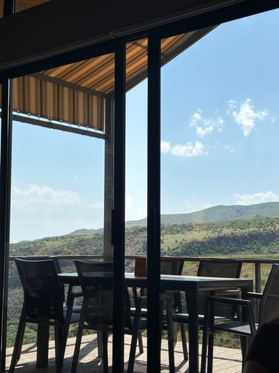 The image captures a serene outdoor dining area with a panoramic view of rolling hills under a bright blue sky. Through a glass partition, a table and chairs are visible on a wooden deck. The chairs are made of dark metal and mesh, and the table appears to be dark metal as well. An awning with striped fabric stretches overhead, offering shade. The landscape beyond is characterized by lush greenery and the rugged texture of the hills. The scene evokes a sense of tranquility and natural beauty, suggesting a peaceful setting for a meal.