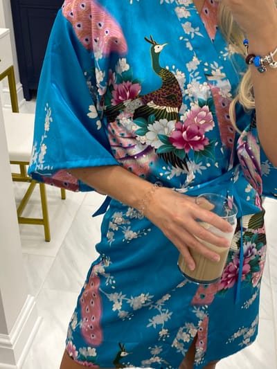 A person is depicted from the waist up, dressed in a bright blue satin robe or kimono adorned with a repeating pattern of peacocks and flowering branches in shades of pink, purple, green, and white. The robe is secured by a matching blue sash at the waist. The individual has blonde hair visible on the right side of the frame. On the right wrist, a silver charm bracelet with blue, purple, and silver charms is present. On the left wrist, a clear, possibly elastic, bracelet with small circular elements is visible. The person holds a clear glass containing a layered light brown beverage, likely a coffee or latte, with the left hand. The setting is an indoor residential or private space within Ridgefield, United States, featuring a light-colored tiled floor, white walls, and partially visible furniture including a dark blue cabinet with gold hardware and a white stool with a gold-colored frame.
