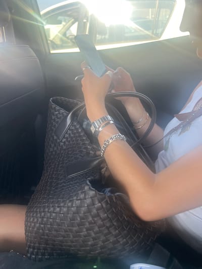 The image displays the interior of a vehicle in Parikia, Greece, under bright daylight conditions, with strong sunlight causing significant lens flare from a side window. A female individual is seated, viewed from the upper torso, engaged with a smartphone held vertically in both hands. She wears a white ribbed tank top. On her left wrist, a silver-toned rectangular watch and a prominent silver-toned curb-link bracelet are visible, along with a thin ring on her left index finger. Her right wrist displays at least two thin gold-toned bracelets. A long necklace with a green pendant is visible on her chest. A large, dark-colored woven tote bag rests in front of her. The vehicle's interior features dark-colored upholstery.