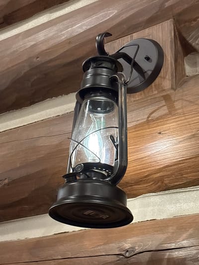 A black, kerosene-style lantern with a glass globe is mounted on a rustic wooden wall. The lantern's light bulb is illuminated. The wall is constructed from horizontally-placed, rough-hewn logs. The scene is indoors.