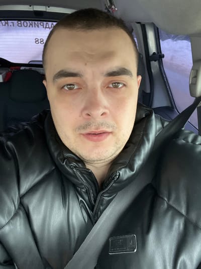 A male individual is depicted in a close-up, front-facing portrait, seated inside a vehicle. He possesses short, dark hair, brown eyes, and light stubble on his chin and upper lip. He is wearing a black, quilted, high-collar jacket, and a gray seatbelt is secured across his chest. In the background, visible through the vehicle's rear window, is reversed Cyrillic text and the number "88" adhered to the glass. To the right, an indistinct, blurry outdoor environment, possibly snowy or overcast, is visible through a side window. A phone mount or similar accessory is attached to the vehicle's interior pillar on the right. The image provides no specific visual cues to determine the exact location within the city of Rogachovo, Russia.