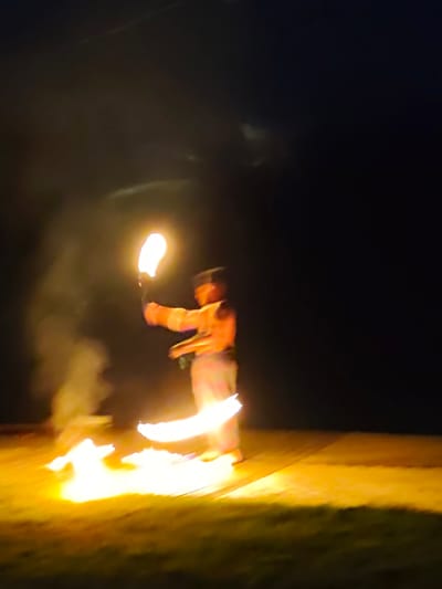 A person is spinning a flaming object in the dark, creating a bright, glowing light. The background is entirely black, suggesting it is nighttime. The foreground appears to be a wooden surface illuminated by the flames. Wisps of smoke are visible rising from the fire. The person is wearing brightly colored clothing, possibly an orange and white striped shirt and white pants. The scene takes place in Bukovik, Montenegro.