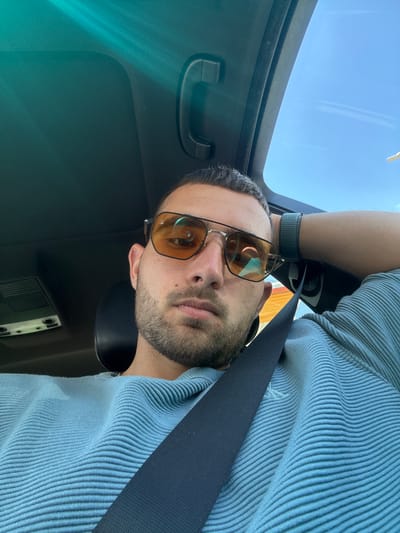 In Polsko Padarevo, Bulgaria, a man with short, dark hair and stubble is seated in a car, looking at the camera. He is wearing orange-tinted sunglasses and a light blue, ribbed long-sleeved shirt. A black seatbelt is fastened across his chest. His right arm is bent, with his hand resting on the back of his head. The car's sunroof is open, revealing a clear blue sky with faint clouds. The interior of the car, including the headliner and a grab handle, is visible.