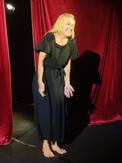 A woman with blonde hair stands barefoot in front of red curtains. She is wearing a black dress with a tie at the waist. Her hands are outstretched and she is smiling. The location is a stage in Bedulu, Indonesia.