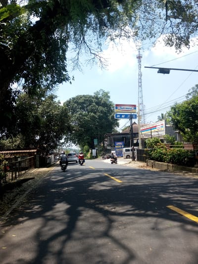 A daytime shot captures a road in a suburban setting. Three motorcyclists travel along the asphalt road, marked with intermittent yellow lines. The road surface is obscured by dappled shadows, likely cast by the dense foliage of the trees lining the roadside. Buildings and signage, including an "Indomaret" sign, are visible on the right side of the road. A tall telecommunications tower rises in the background, reaching above the buildings and trees. The location appears to be Nawng Hkar, Myanmar, based on the language on the Indomaret sign.