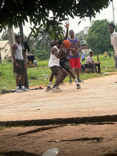In Amansee, Nigeria, a group of young men are engaged in a basketball game on an outdoor court. One man, wearing a grey tank top and black shorts, stands on the left side of the court. Another man in a white tank top and black shorts is positioned in the center, holding a basketball. To his right, a third man in a purple jersey with a graphic print and red shorts is visible. Several other individuals are present in the background, some observing the game, while others appear to be walking or standing in the grassy area surrounding the court. A blue and yellow motorized tricycle is parked behind the players. The court itself is made of a light-colored, possibly concrete, surface with visible lines. The surrounding environment is characterized by lush green grass, trees, and some scattered debris. A building with a light-colored roof and windows is situated in the upper left portion of the image. Overhead, tree branches and leaves partially obscure the sky.