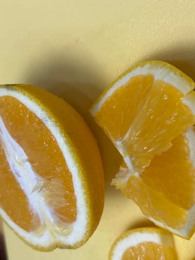 The image shows a close-up view of cut oranges on a pale yellow surface. On the left side of the frame is half of an orange, showing its segmented interior and thick rind. To the right, there are several wedges of orange, some overlapping, with their juicy flesh and white pith clearly visible. The lighting is bright, highlighting the vibrant colors of the fruit. There are no people, animals, or specific urban features visible that would indicate a location in Засечное, Russia.