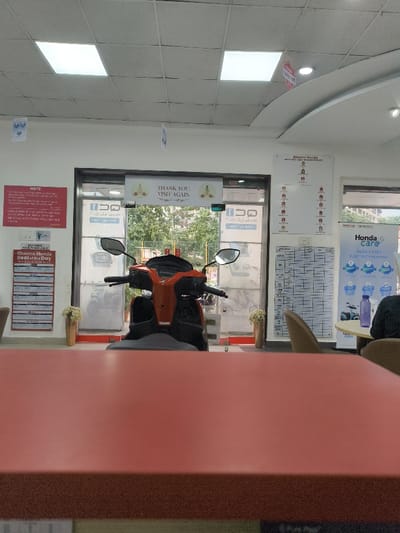 The image shows a Honda dealership in Navi Mumbai, India. The image was taken inside the dealership. A red Honda scooter is displayed in the center of the image, behind a red desk. There is a glass door to the left, a white wall with a "Thank You, Visit Again" sign above a large whiteboard, and a wall with the dealership's logo to the right of the scooter. The floor is tiled. The scene appears to be taken during the day. There are multiple potted plants around the dealership. In the right foreground, a beige chair is visible, while in the right background a table is occupied by someone. There is a sign hanging above the table for "Honda Care" and "Hassle-free Electric Freedom."