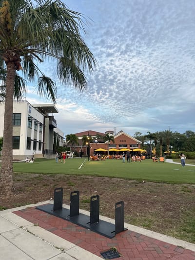The image depicts an outdoor scene in Tampa, Florida, United States. The sky is partly cloudy. A tall palm tree is on the left side. A modern, multi-story building with large windows and a flat roof is located in the lower center. A red brick building with a peaked roof and yellow umbrellas shading outdoor seating is in the background. A grassy area is visible in front of these buildings, with people walking and seated at tables. In the foreground, there are three black metal structures, possibly bike racks, situated on a paved area with brick accents.