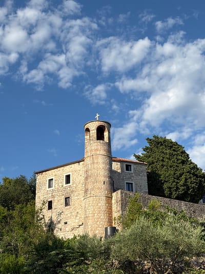 A stone church with a bell tower topped by a cross is nestled among lush green trees under a bright blue sky with scattered white clouds. The church features a cylindrical bell tower with arched openings and a small cross at its apex. The main body of the church is constructed of rough-hewn stone blocks and has several rectangular windows. A stone wall borders the property, and dense foliage surrounds the building. The image appears to be taken from a lower vantage point, looking up towards the church, which is situated on a hillside in the region of Podostrog, Montenegro. No people are visible in the image.