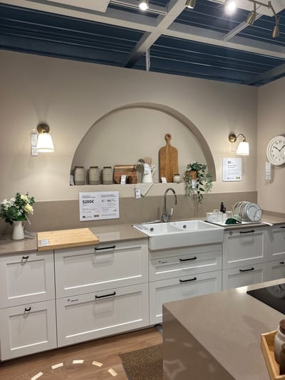 This is an image of a kitchen display in a retail store, likely IKEA, in Siero, Spain. The scene depicts a white farmhouse sink with a chrome faucet, set into a light brown countertop. To the left of the sink, there is a white pitcher with a bouquet of white flowers and greenery. A wooden cutting board is placed on the countertop in front of the pitcher.

Above the sink, within an arched alcove, are three beige ceramic jars with brown lids. To the right of the jars is a wooden cutting board leaning against the wall. Further to the right, a potted plant with green leaves hangs from the wall.

The cabinetry below the countertop is white with black handles. Several price tags are visible throughout the display. One tag on the left side of the image indicates a price of 19.99. Another larger sign on the countertop to the left of the sink states "Precio de esta combinación de cocina: 5200€" and "Disfrútala hoy y págala cómodamente en 48 meses" with additional smaller text and an IKEA logo. A square white sign with QR code and text is mounted on the wall to the right of the hanging plant.

On the far right of the image, a round white clock with black numerals is mounted on the wall. Below the clock, a white tag with the price 19.99 is hanging. A dish drying rack containing white plates and green bowls is situated on the countertop to the right of the sink. The environment appears to be a brightly lit showroom with a beige-colored wall and a blue ceiling structure visible at the top.