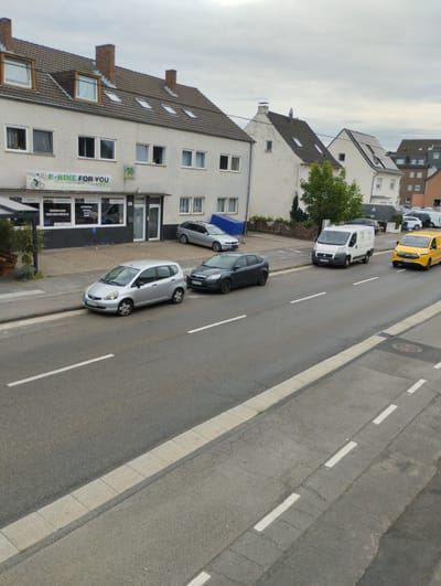 This is an eye-level, medium shot of a street in Troisdorf, Germany. The scene is captured on an overcast day, with a cloudy sky filling the upper portion of the frame. In the foreground, a paved road with white lane markings dominates the lower half of the image. To the left, a sidewalk separates the road from a commercial building.

The building appears to be a shop with a large sign that reads "E-BIKE FOR YOU." It has multiple windows and doorways. In front of the shop, a silver hatchback car is parked. Further down the street, a dark-colored sedan is also parked.

Several vehicles are in motion on the road. A white panel van is driving away from the camera, followed by a yellow panel van. In the background, more buildings and parked cars are visible. The overall environment suggests a typical urban street scene with commercial activity.
