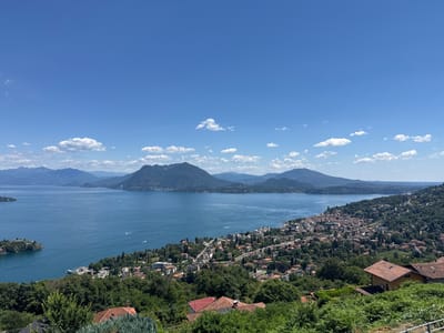 The image depicts a panoramic view of Lake Maggiore, with the city of Someraro, Italy, nestled along its shores. The lake is a vibrant blue, dotted with a few small boats. Mountains form a majestic backdrop to the scene, their peaks shrouded in a hazy blue. The city itself is a tapestry of terracotta-tiled roofs and whitewashed buildings, interspersed with lush greenery. A prominent feature is a small island located off the coast, which appears to be a fortified settlement. The foreground is dominated by dense foliage and the rooftops of houses, suggesting a vantage point from a hillside overlooking the city and the lake. The sky is a clear, brilliant blue, adorned with a few fluffy white clouds. The scene is bathed in bright daylight, indicating a sunny day.
