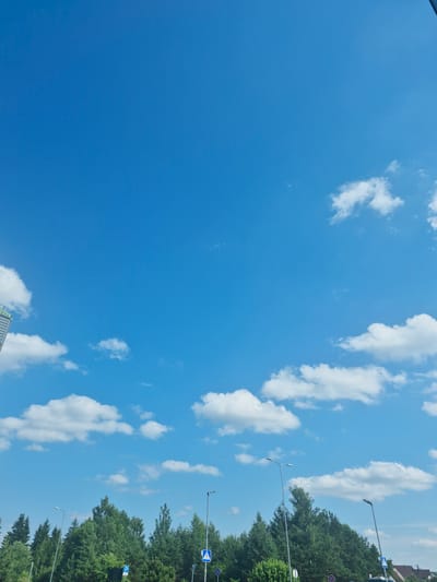 The image displays a bright blue sky with scattered white clouds. On the right side of the frame, a line of lush green trees is visible, suggesting an outdoor setting. Several streetlights with modern designs are interspersed along the tree line, indicating an urban or suburban environment. A pedestrian crossing sign, a common road marking, is also present. Further down the line, a parking sign is visible. The overall impression is of a clear, sunny day. The orientation of the image is landscape, with the sky occupying most of the frame. The presence of road signs and streetlights suggests a public area, possibly a street or a road in Pabradė, Lithuania. There are no people visible in the image, and the focus is on the natural elements and urban infrastructure. The time of day is likely daytime, given the bright sky and sunlight illuminating the scene.