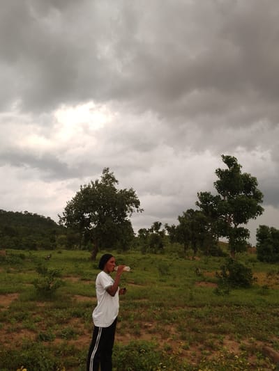 A young woman stands in a field of green grass and scattered trees, drinking from a bottle. She wears a white t-shirt, black pants with white stripes, and a black headscarf. The sky above is filled with dramatic, dark clouds. The landscape is gently rolling, with trees dotting the hills in the background. The area appears to be rural, likely in or around the city of Balewa, Nigeria, given its characteristic vegetation and atmospheric conditions. The image captures a moment of pause amidst what appears to be an impending storm.