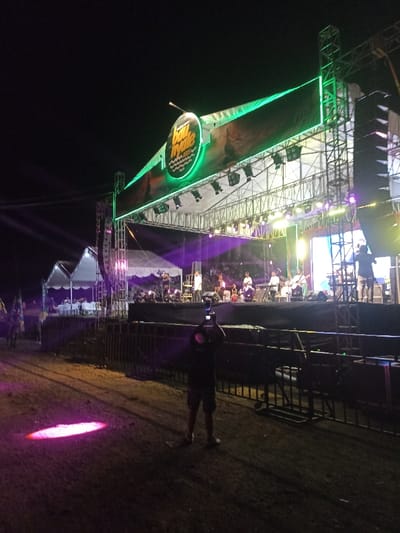 The image captures an outdoor music concert at night in Kecamatan Pujut, Indonesia. A large, illuminated stage dominates the scene, constructed from metal trussing and adorned with a prominent green-lit banner. The banner features a circular logo with the text "BAU NYALE 2023".
On the stage, a band performs, with several musicians visible playing instruments such as guitars, keyboards, and drums, under a wash of purple and yellow stage lights. Speaker stacks flank the stage, and white tents are set up to the left of the performance area.
In the foreground, a person stands facing the stage, holding up a smartphone or camera to record the event. Metal crowd control barriers separate the audience area from the stage. A bright pink spotlight casts an oval shape on the dark ground directly in front of the foreground observer. The dark sky indicates it is nighttime, and the artificial lighting provides the primary illumination for the event.