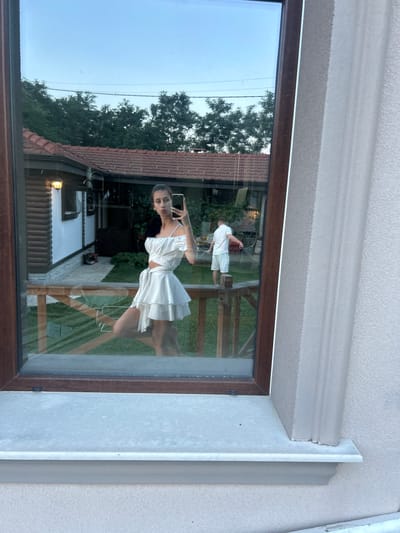 A young woman in a white two-piece outfit, consisting of a cropped top and a tiered skirt, is taking a selfie in a reflection on a window. She is standing with her left leg bent and resting on a wooden railing, while her right leg is extended. In the reflection, a man in a white t-shirt and shorts is visible watering the grass with a hose in the background. The scene appears to be a residential area, possibly a backyard, with a house with a red tile roof and greenery visible. The location is stated to be Войводино, Bulgaria.